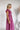 Side view of the Living In A Dream Maxi Dress that features a pink colored material, a V neckline, a short ruffle sleeve, a knot detail at the bust, an open back with adjustable tie closures, and a maxi length.
