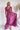 Frontal view of the Living In A Dream Maxi Dress that features a pink colored material, a V neckline, a short ruffle sleeve, a knot detail at the bust, an open back with adjustable tie closures, and a maxi length.