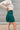 Side view of the In Harmony Skirt that features a hunter green material, a high-rise fit, a thick waist band, a pleated design throughout, and a back zipper closure