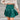 Frontal view of the In Harmony Skirt that features a hunter green material, a high-rise fit, a thick waist band, a pleated design throughout, and a back zipper closure