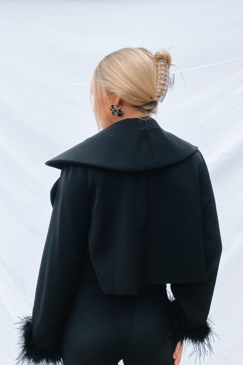 Back view of the The Always The Optimist Crop Blazer that features black fabric, an open front with a collar and lapel, a long sleeve with feather trim, and a cropped fit