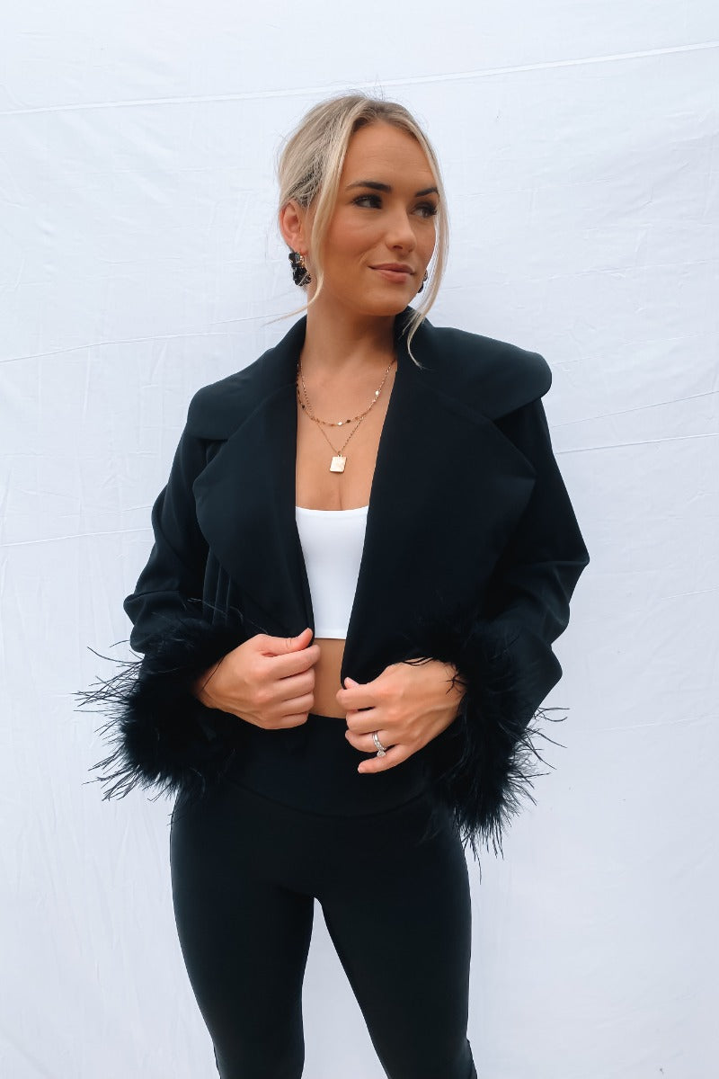 Frontal view of the The Always The Optimist Crop Blazer that features black fabric, an open front with a collar and lapel, a long sleeve with feather trim, and a cropped fit