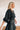 Side view of the Show Time Bodysuit that features a black satin material, a V neckline, a flowy half sleeve with a feather trim, and a bikini bottom with a snap closure.