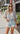 Front view of model wearing the Love Like This Bodysuit, which has an ivory and grey knit material with a floral print, a V surplice neckline, an oversized half sleeve, and a bikini bottom with a snap closure. Worn with denim shorts.