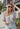 Front view of model wearing the Love Like This Bodysuit, which has an ivory and grey knit material with a floral print, a V surplice neckline, an oversized half sleeve, and a bikini bottom with a snap closure. Worn with denim shorts.