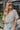 Front view of model wearing the Love Like This Bodysuit, which has an ivory and grey knit material with a floral print, a V surplice neckline, an oversized half sleeve, and a bikini bottom with a snap closure. Worn with denim shorts.