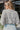 Back view of model wearing the Love Like This Bodysuit, which has an ivory and grey knit material with a floral print, a V surplice neckline, an oversized half sleeve, and a bikini bottom with a snap closure. Worn with denim shorts.
