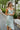 
Frontal view of the Endless Nights Midi Dress that features a white silk material, a sage green swirl print, a cowl neckline, a sleeveless design with thin adjustable straps, a side slit, and a midi length.
