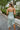 Back view of the Endless Nights Midi Dress that features a white silk material, a sage green swirl print, a cowl neckline, a sleeveless design with thin adjustable straps, a side slit, and a midi length.
