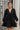 Frontal view of the Line Of Sight Dress In Black that features a black colored material, a round notched neckline with adjustable ties, long sleeves, vertical ruffle details, and a flowy fit.