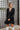 Frontal view of the Line Of Sight Dress In Black that features a black colored material, a round notched neckline with adjustable ties, long sleeves, vertical ruffle details, and a flowy fit.