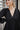 Frontal view of the Line Of Sight Dress In Black that features a black colored material, a round notched neckline with adjustable ties, long sleeves, vertical ruffle details, and a flowy fit.