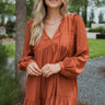 Frontal view of the Line Of Sight Dress In Rust that features a rust colored material, a round notched neckline with adjustable ties, long sleeves, vertical ruffle details, and a flowy fit.