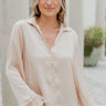 Frontal view of the On To The Next Top that features a champagne satin material, a collar neckline, a long sleeve with feather detailing at the cuff, a button-up front, and a flowy fit.