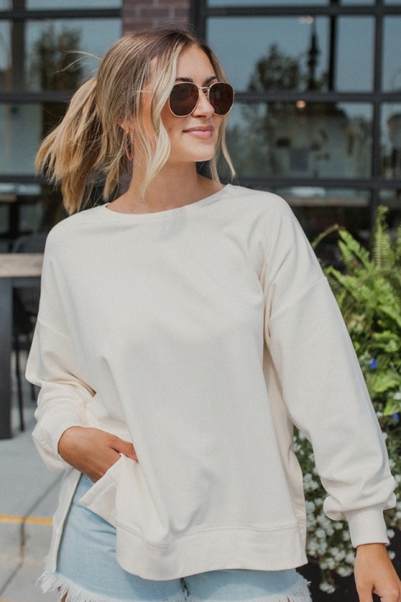 Frontal view of the Where To Next Top that features a soft cream material, a round neckline, a long cuffed sleeve, two side slits, and a thick bottom hem.