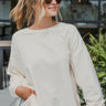 Frontal view of the Where To Next Top that features a soft cream material, a round neckline, a long cuffed sleeve, two side slits, and a thick bottom hem.