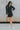 Full body front view of model wearing the Talk The Talk Dress in Black that has washed black denim fabric, mini length, a frayed hem, a hidden quarter button up, a collared neck, a front chest pocket and long sleeves with cuffs