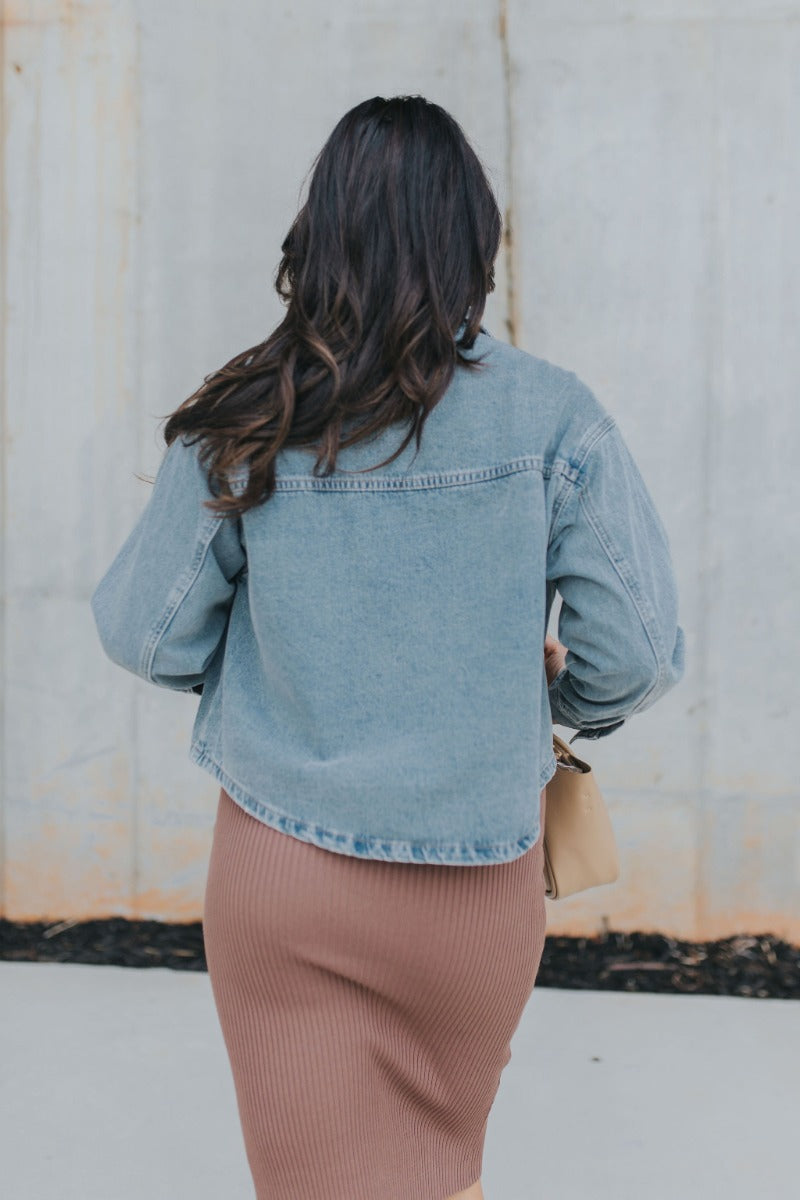 Back view of model wearing the Good Times Denim Jacket that has medium wash denim fabric, distressed details, pockets, silver snaps, a collared neck, and long sleeves with buttoned cuffs