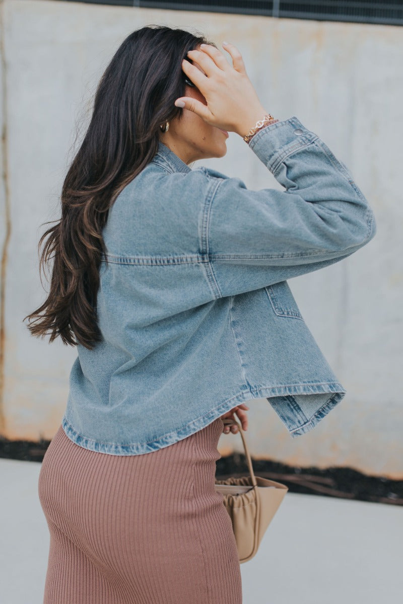 Side view of model wearing the Good Times Denim Jacket that has medium wash denim fabric, distressed details, pockets, silver snaps, a collared neck, and long sleeves with buttoned cuffs