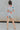 Full body side view of model wearing the Dreamy Floral Romper which features white, red, tan, sage, pink, muted yellow and blue cotton fabric, floral pattern, folded hem, two side pockets, two back pockets, button snap closure, two front chest pockets, co
