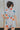 Back view of model wearing the Dreamy Floral Romper which features white, red, tan, sage, pink, muted yellow and blue cotton fabric, floral pattern, folded hem, two side pockets, two back pockets, button snap closure, two front chest pockets, collared nec