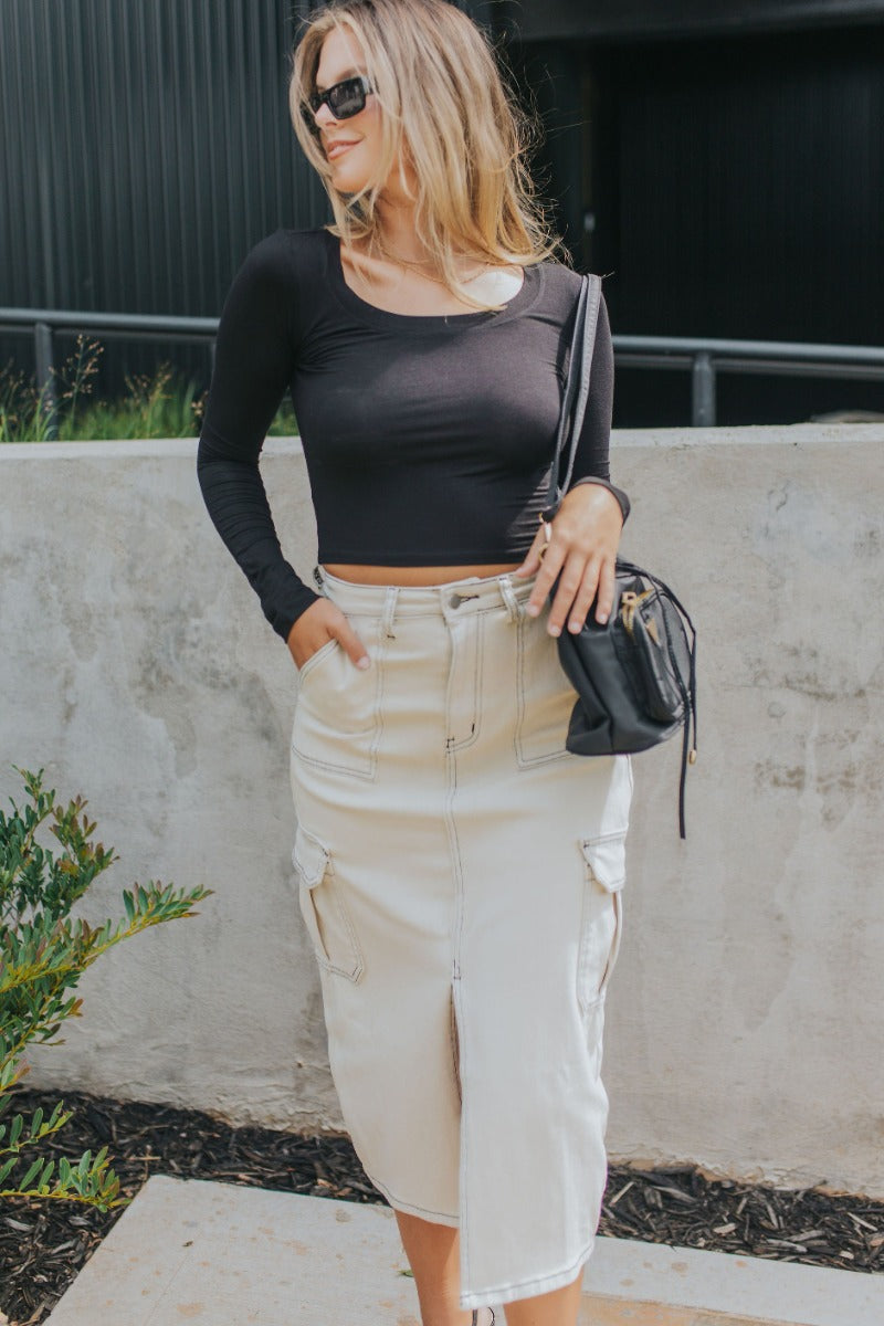 front view of model wearing the Near Or Far Cargo Midi Skirt that has cream denim fabric, a front slit, black stitching, a front zipper, pockets, and belt loops.