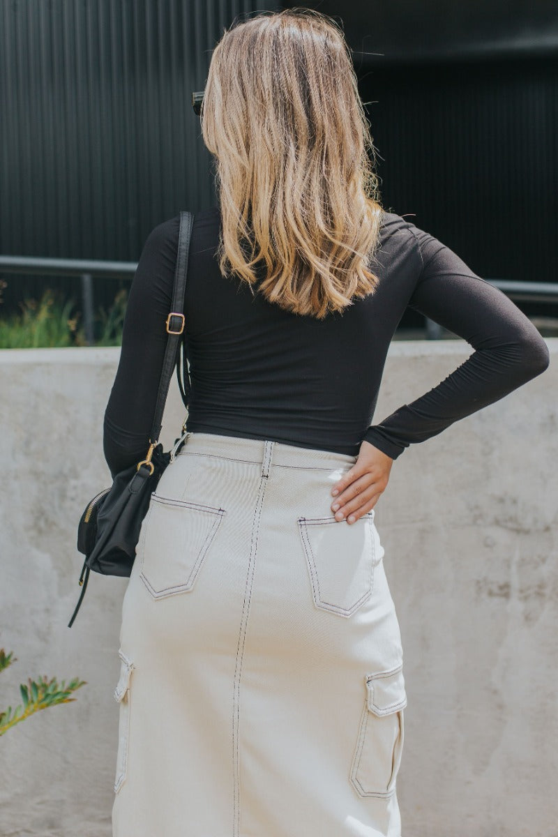 Upper back view of model wearing the Near Or Far Cargo Midi Skirt that has cream denim fabric, a front slit, black stitching, a front zipper, pockets, and belt loops.