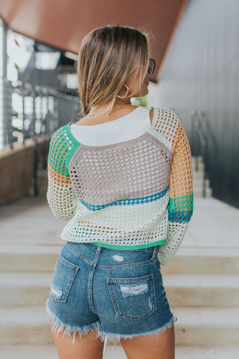 Back view of model wearing the Let It Be Sweater in Mint which features green, taupe, peach, blue and mint green open knit fabric, cropped waist, round neckline and long flare sleeves.