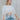 Front view of model wearing the Anywhere With You Top which features off white knit fabric, cropped waist, tassel hem details, open knit details, round neckline with monochrome stitch details and short sleeves with tassel hem details.