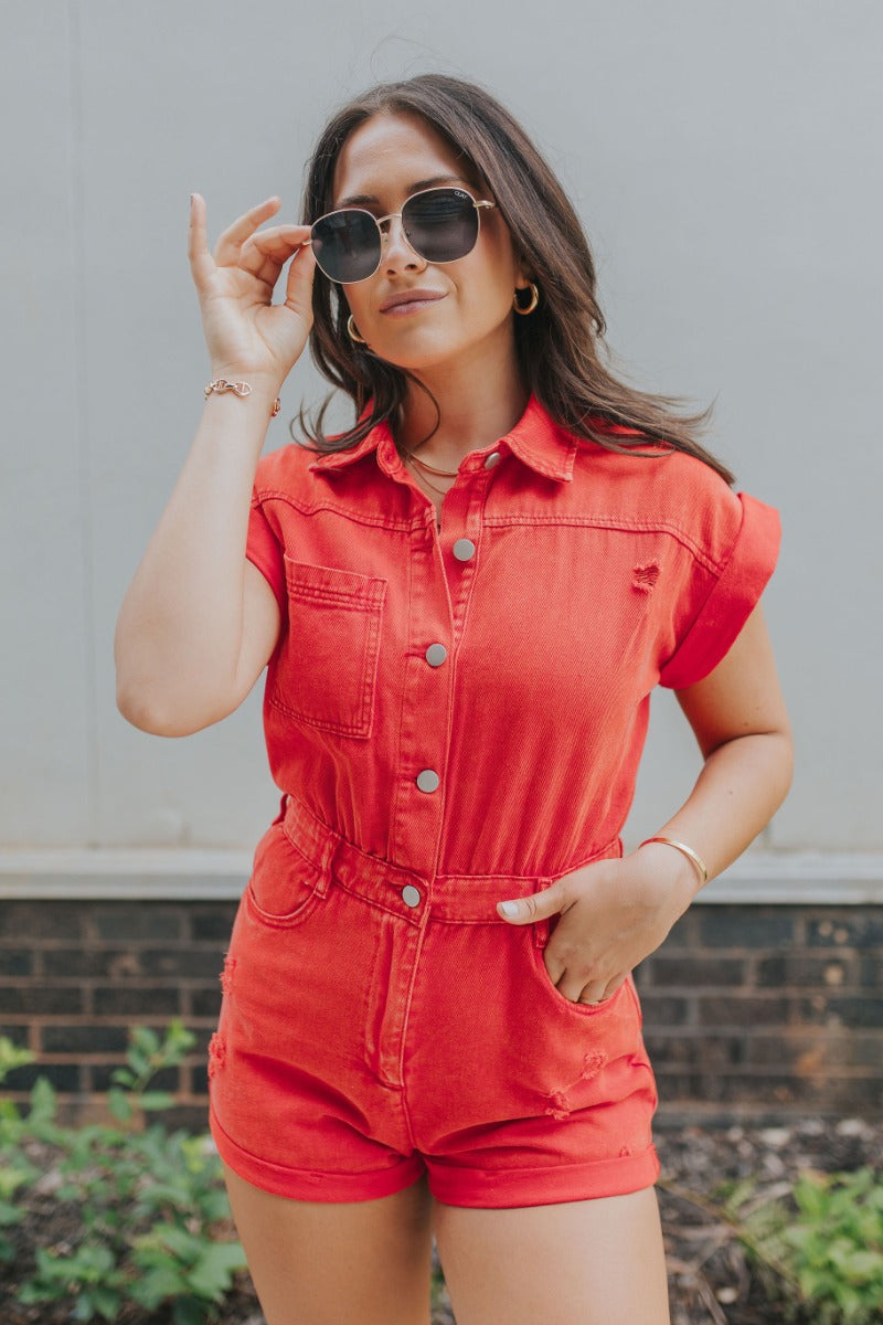 Front view of model wearing the Game On Romper which features washed red denim fabric, distressed details, two front pockets, one chest pocket, folded hem shorts, an elastic waistband in the back, a hidden zipper and button up closure, a collared neckline