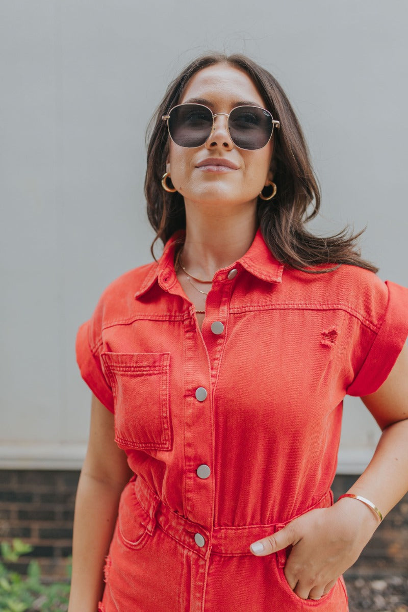 Front close up view of model wearing the Game On Romper which features washed red denim fabric, distressed details, two front pockets, one chest pocket, folded hem shorts, an elastic waistband in the back, a hidden zipper and button up closure, a collared