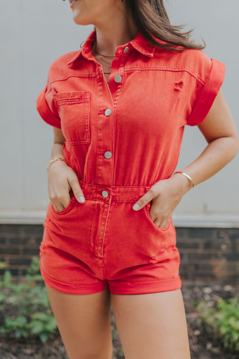 Front close up view of model wearing the Game On Romper which features washed red denim fabric, distressed details, two front pockets, one chest pocket, folded hem shorts, an elastic waistband in the back, a hidden zipper and button up closure, a collared
