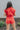 Back view of model wearing the Game On Romper which features washed red denim fabric, distressed details, two front pockets, one chest pocket, folded hem shorts, an elastic waistband in the back, a hidden zipper and button up closure, a collared neckline,