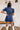 Back view of the Running The Show Denim Romper that features a navy colored denim with some stretch, a surplice neckline that snaps with a collar, half-length sleeves, a front zipper, pockets, pleating on the legs, and a 3" inseam.