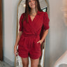 Frontal view of the  Running The Show Denim Romper that features a crimson colored denim with some stretch, a surplice neckline that snaps with a collar, half-length sleeves, a front zipper, pockets, pleating on the legs, and a 3" inseam.