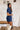 Side view of the Running The Show Denim Romper that features a navy colored denim with some stretch, a surplice neckline that snaps with a collar, half-length sleeves, a front zipper, pockets, pleating on the legs, and a 3" inseam.