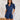 Frontal view of the Running The Show Denim Romper that features a navy colored denim with some stretch, a surplice neckline that snaps with a collar, half-length sleeves, a front zipper, pockets, pleating on the legs, and a 3" inseam.