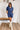 Frontal view of the Running The Show Denim Romper that features a navy colored denim with some stretch, a surplice neckline that snaps with a collar, half-length sleeves, a front zipper, pockets, pleating on the legs, and a 3" inseam.