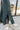 Side view of model wearing The Get In The Groove Pants  features textured dark green fabric with navy stripes, an elastic waist, wide flowy legs, and ankle slits.