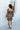 Full body back view of model wearing the I Like Your Vibe Floral Dress, which features a brown chiffon material, a taupe floral print, a curved neckline, long sleeves, a tier design with ruffles, a smocked back, and a mini length
