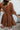 Back view of model wearing the Spice It Up Dress, which features camel-colored fabric, a v-neckline with a cutout, a v-back with a tie, half sleeves, and a flowy fit
