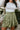 Close-up front view of model wearing the Feeling Lucky Skirt, whcih features green silk fabric with a green lining, a pleated waistband with a smocked back, a front zipper detail, and two front flap pockets