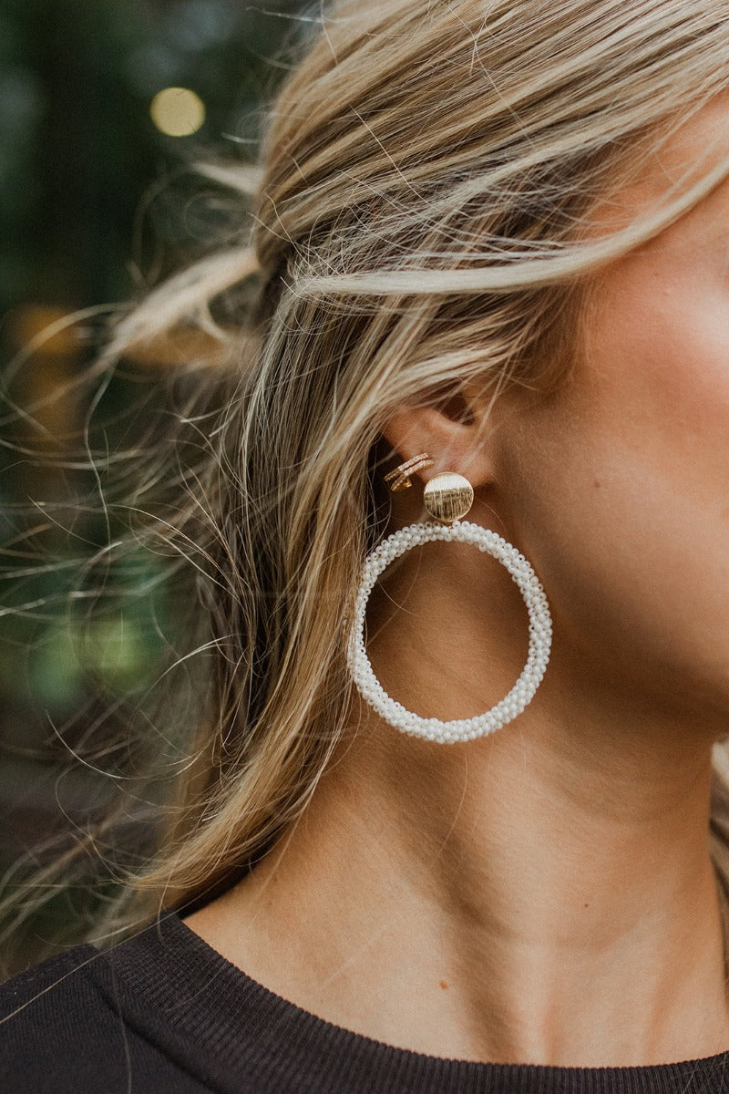 Close up image of model wearing the Upper East Side Earring, which features gold circles on post backs with large circular white beaded pendants.