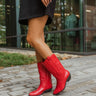 Side view of model wearing The Red Hot Western Boot which features faux leather, western stitching, and a 2" heel. These boots run small, we suggest sizing up half a size! 