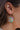 Close up image of model's ear- model is wearing the Snow Day Earrings, that have a gold hexagon pendant with a clear center, which is filled with iridescent multicolor snowflakes.