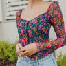 Close up view of model wearing The Slow Down Top which features black mesh fabric with a multi-colored floral print, a lined bodice, a corset front with hook and eye closures, and long sleeves with elastic shoulders.