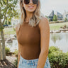 Close front view of model wearing the Take It Easy Bodysuit, which features camel-colored ribbed knit fabric, a round neckline, and a thong bottom with snap closures. Bodysuit is worn with jeans.