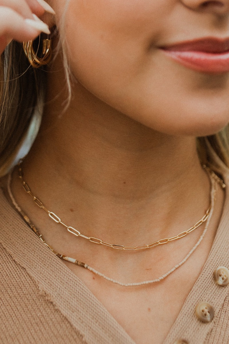 Close view of model wearing the Free To Be Necklace, which features double strands: 1 shorter gold link chain, and 1 strand with beige, brown, and gold beads.