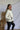 Side view of model wearing the Yosemite Puffer Jacket that has ivory puffer fabric, two front pockets, black zipper details, a zip-up front, and a hood. Worn over black top.