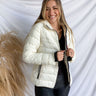 Front view of model wearing the Yosemite Puffer Jacket that has ivory puffer fabric, two front pockets, black zipper details, a zip-up front, and a hood. Worn over black top.